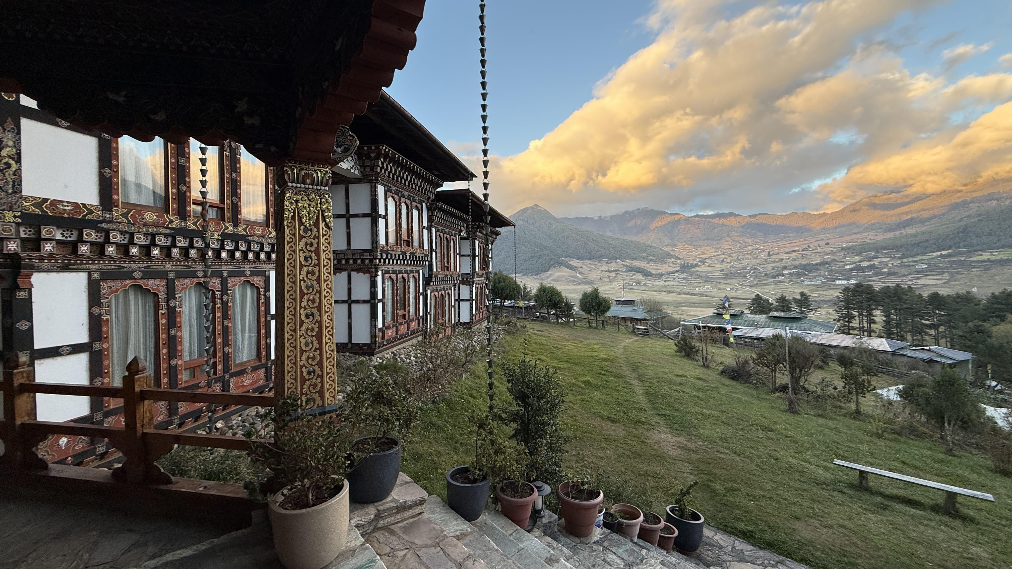 Traditional Bhutanese lodge overlooking Phobjikha Valley at sunset, with carved wooden architecture, prayer flags, and rolling mountain landscape.