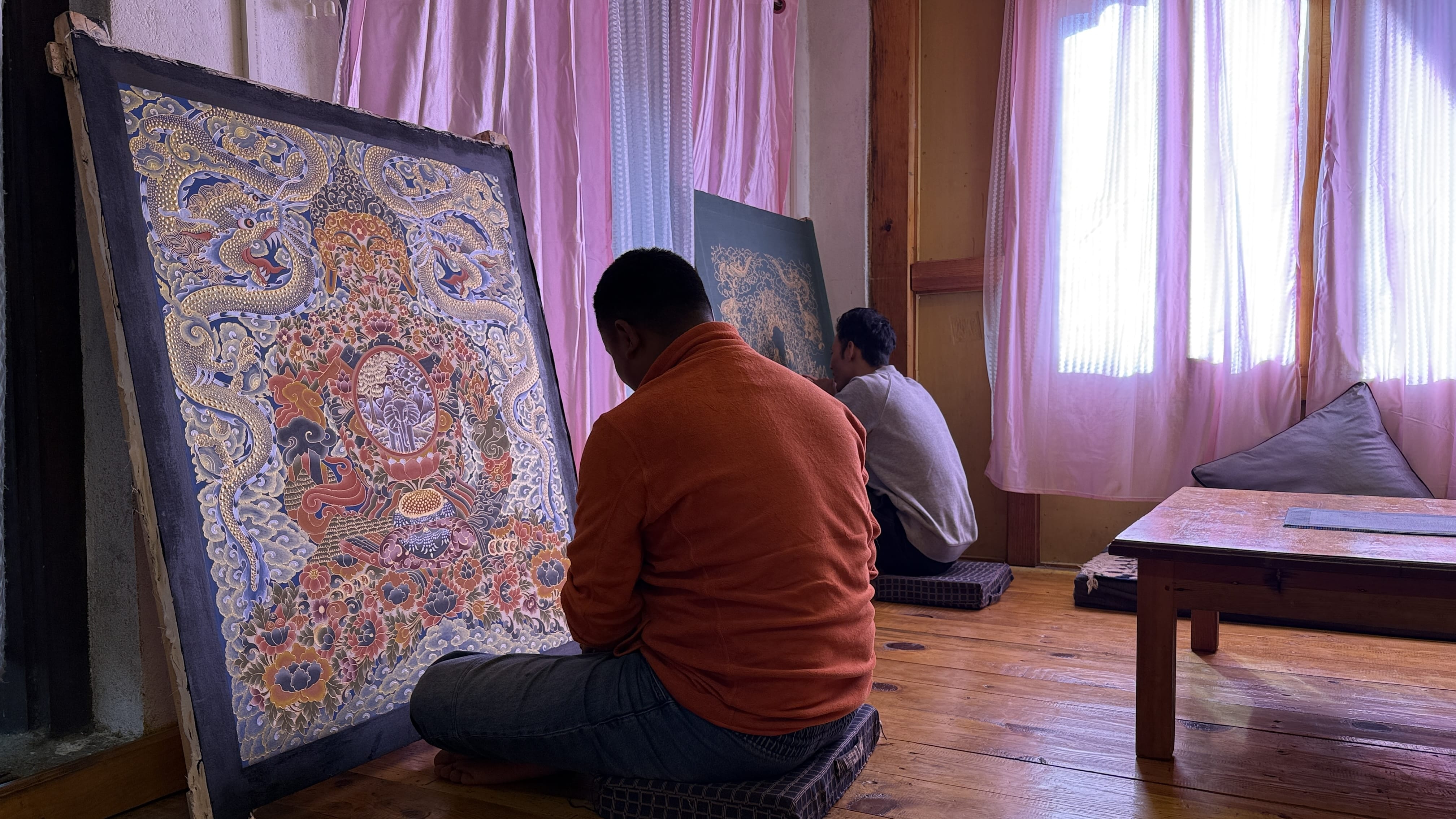 11 Days in Bhutan: thangka artists at work in a small studio near the Divine Madman’s monastery, painting a detailed Buddhist scene on canvas by window light.