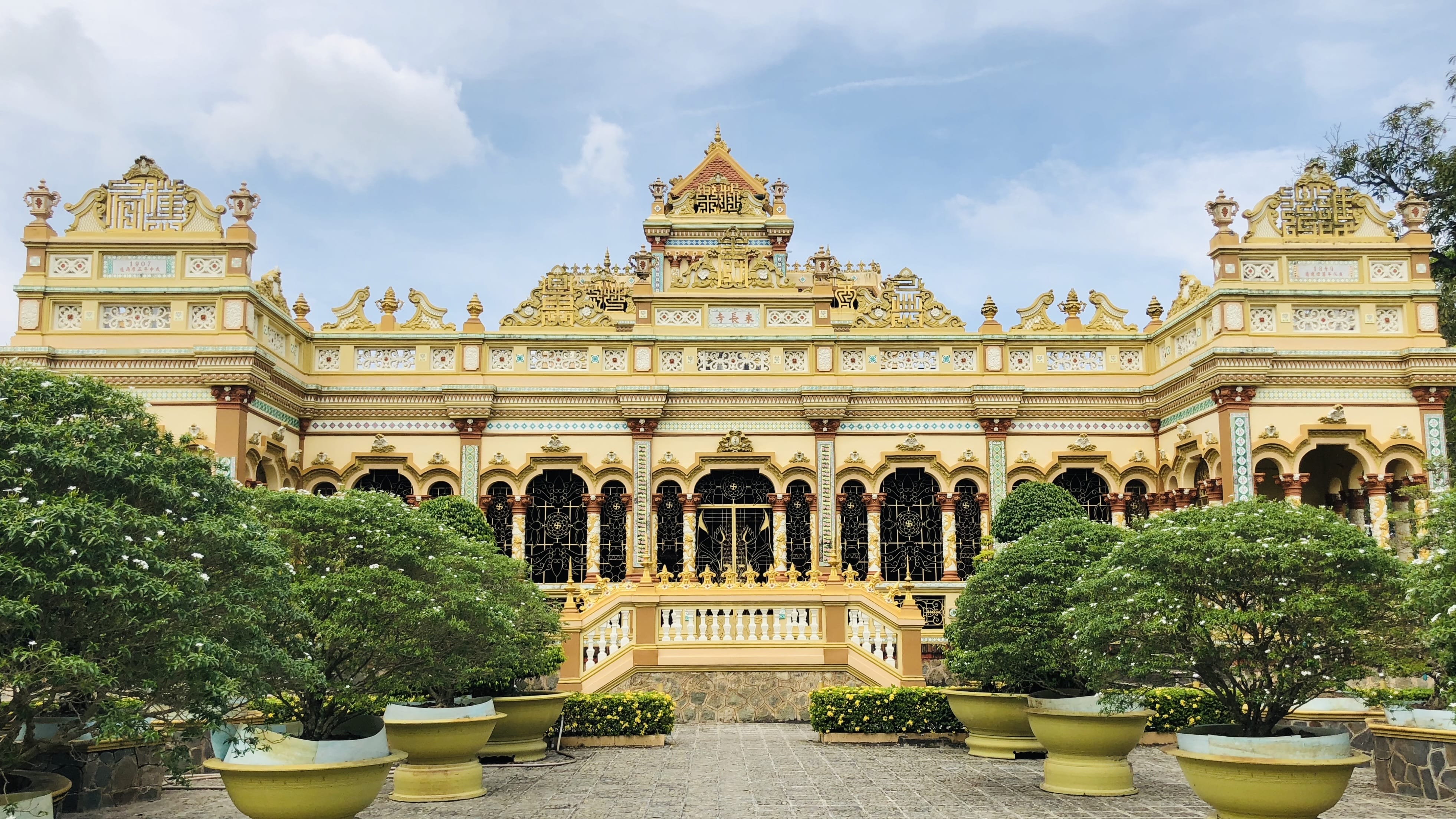 Ornate yellow and gold Vinh Trang Temple with decorative arches, carved details, and large potted bonsai trees in the courtyard under a cloudy sky.