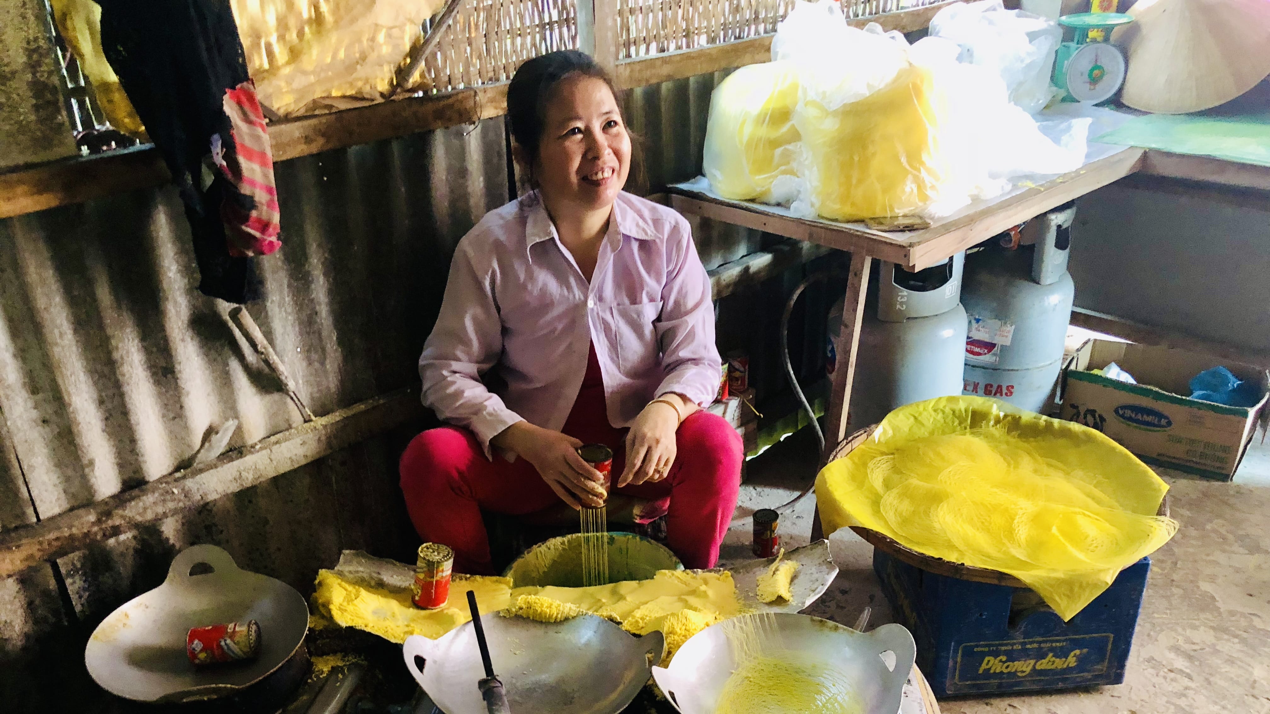 Woman in a Mekong Delta home pressing thick rice batter through a metal mold into a wok to make fresh rice noodles by hand.
