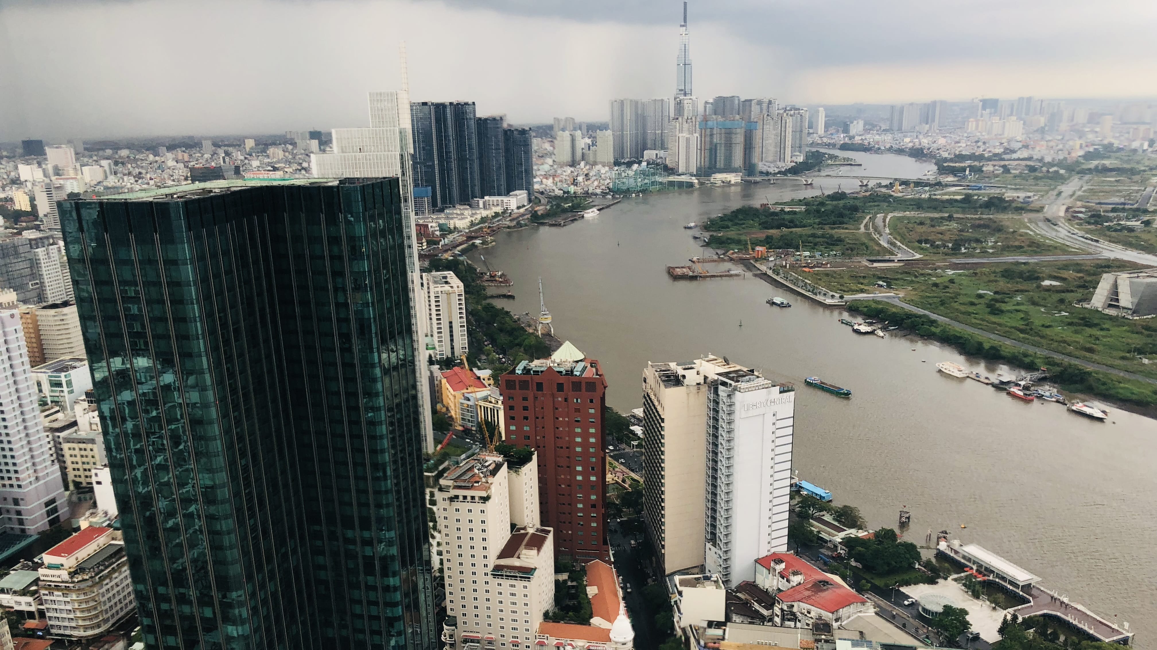 Aerial view of Ho Chi Minh City skyline and Saigon River with modern skyscrapers including Landmark 81 under cloudy skies.