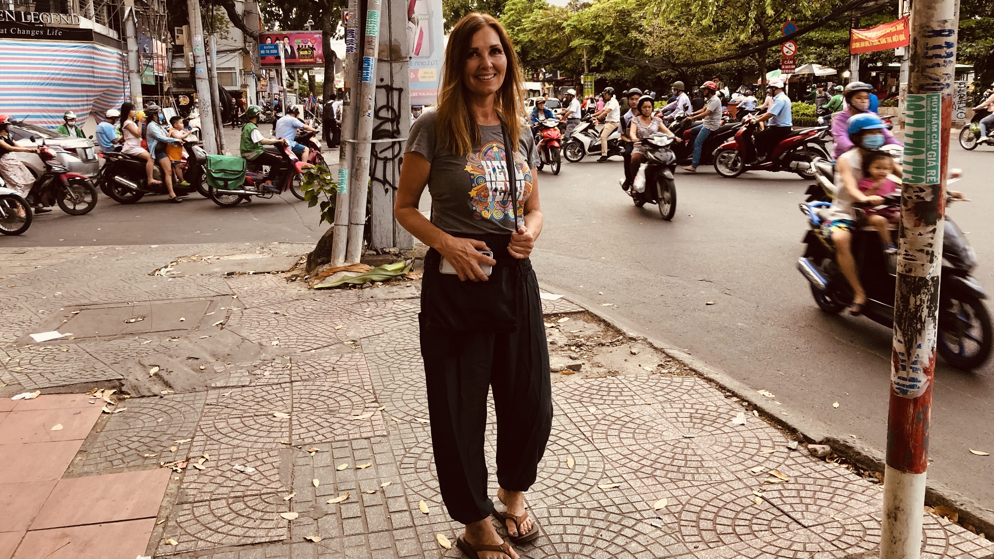 Woman standing on a Ho Chi Minh City sidewalk with heavy motorbike traffic behind her, wearing a graphic T-shirt and loose black pants.